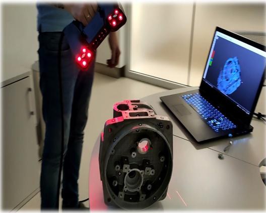 A person using a handheld 3D scanner on a mechanical part, with a laptop displaying the scan data